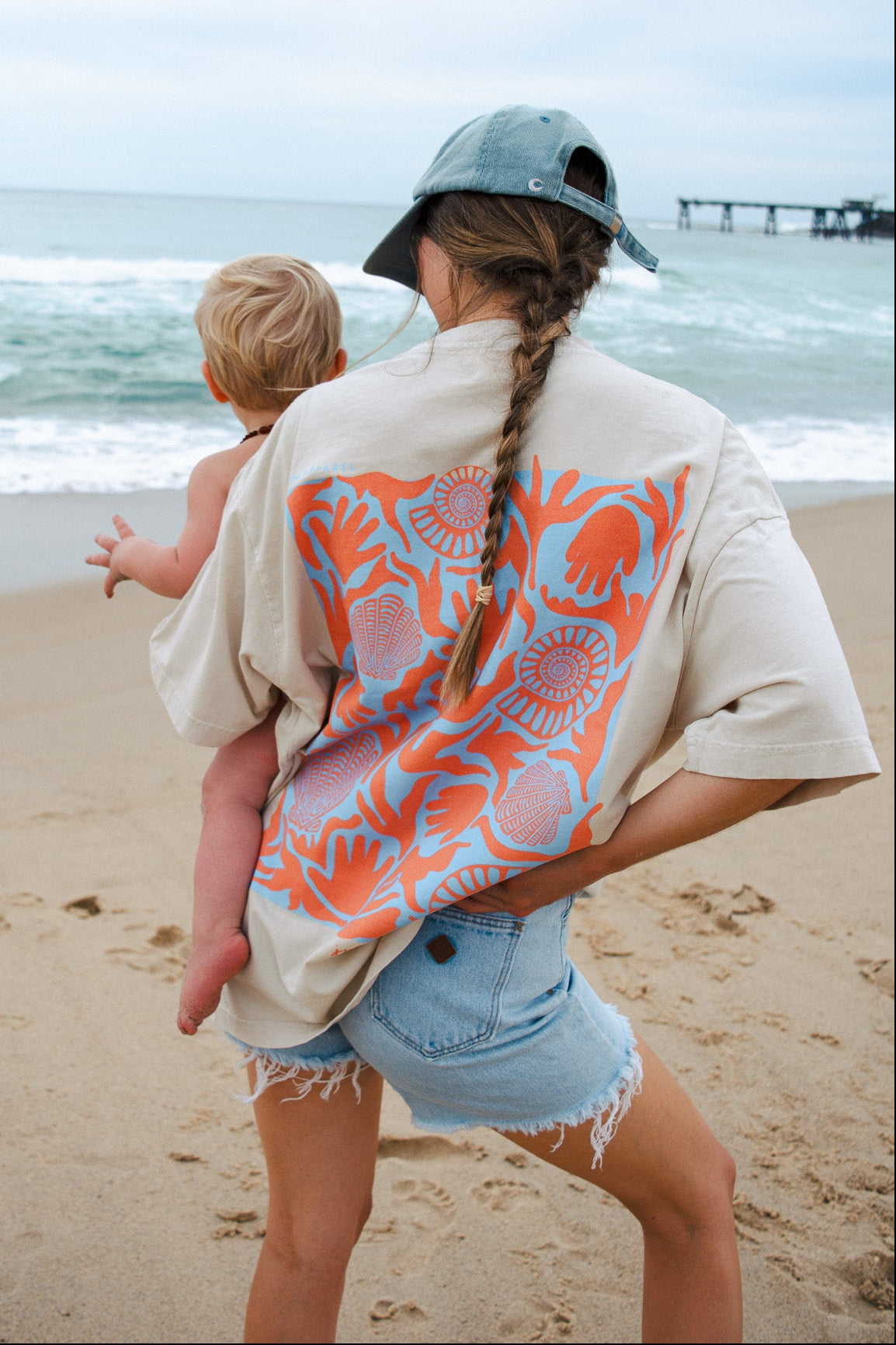 Woman holding a child on a beach with a colorful custom graphic tee in denim shorts.